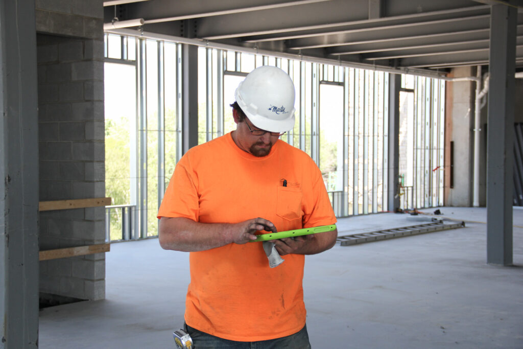 Construction worker in orange shirt uses tablet and digital planning technology to determine where walls will be built in a new hospital.